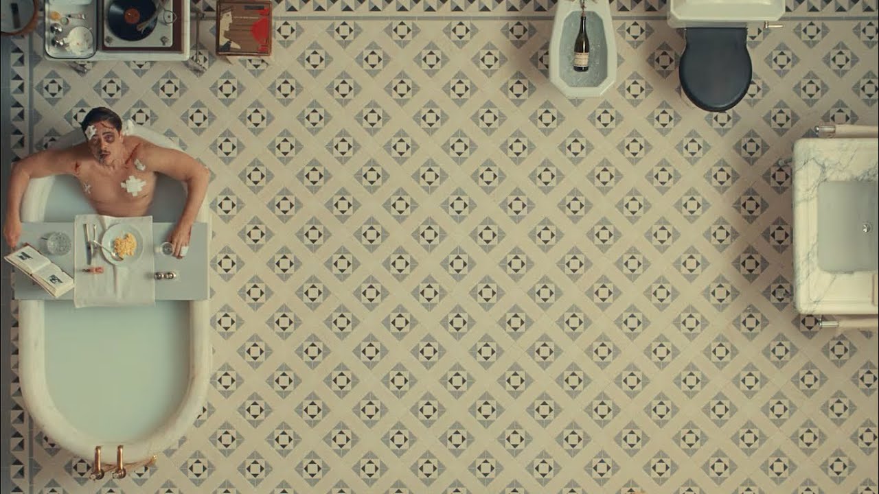 still from The Phoenican Scheme - Benecio del toro looks up from his bath, with the rest of his austere mid-century rich guy bathroom filling the frame